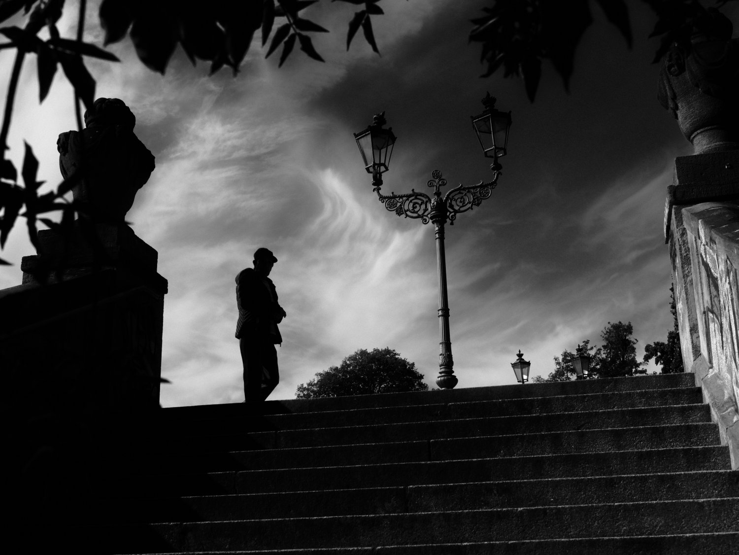 Schwarzweißfoto: Person auf Treppen in Schöneberg mit Straßenlaterne und Statue, dramatischer Himmel